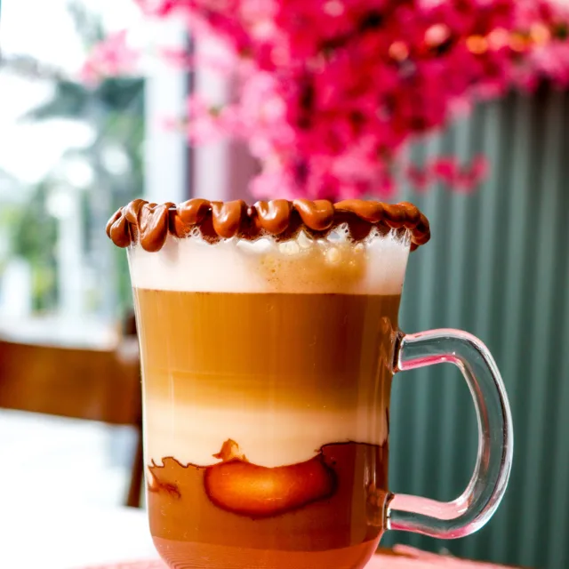 Café em camadas na caneca de vidro, com a borda de chocolate e a árvore florida ao fundo.