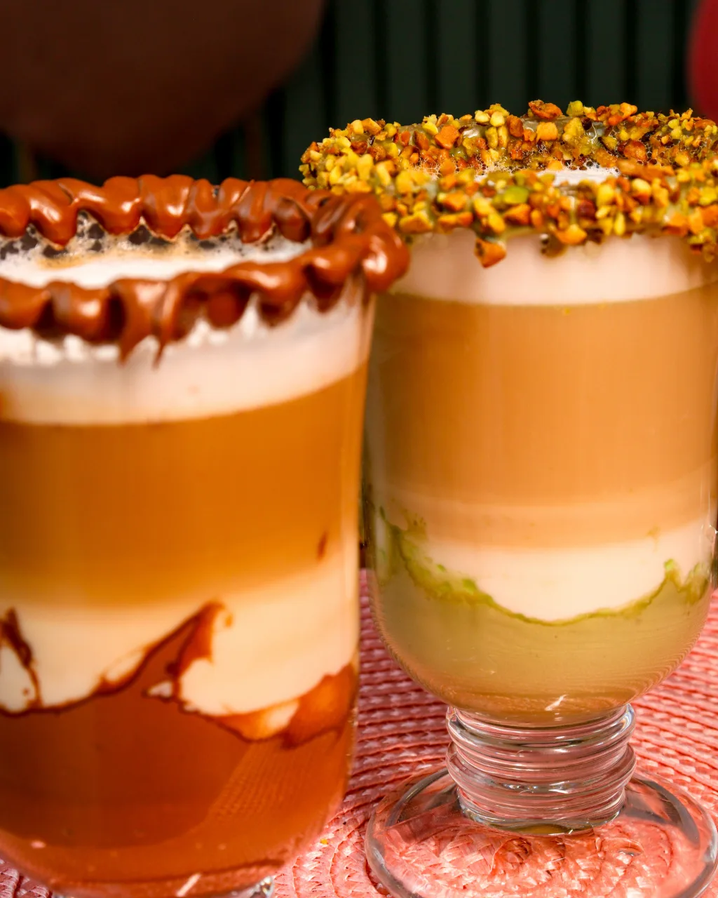 Dois cafés em camadas lado a lado, um com a borda de chocolate e outro com a borda de pistache.