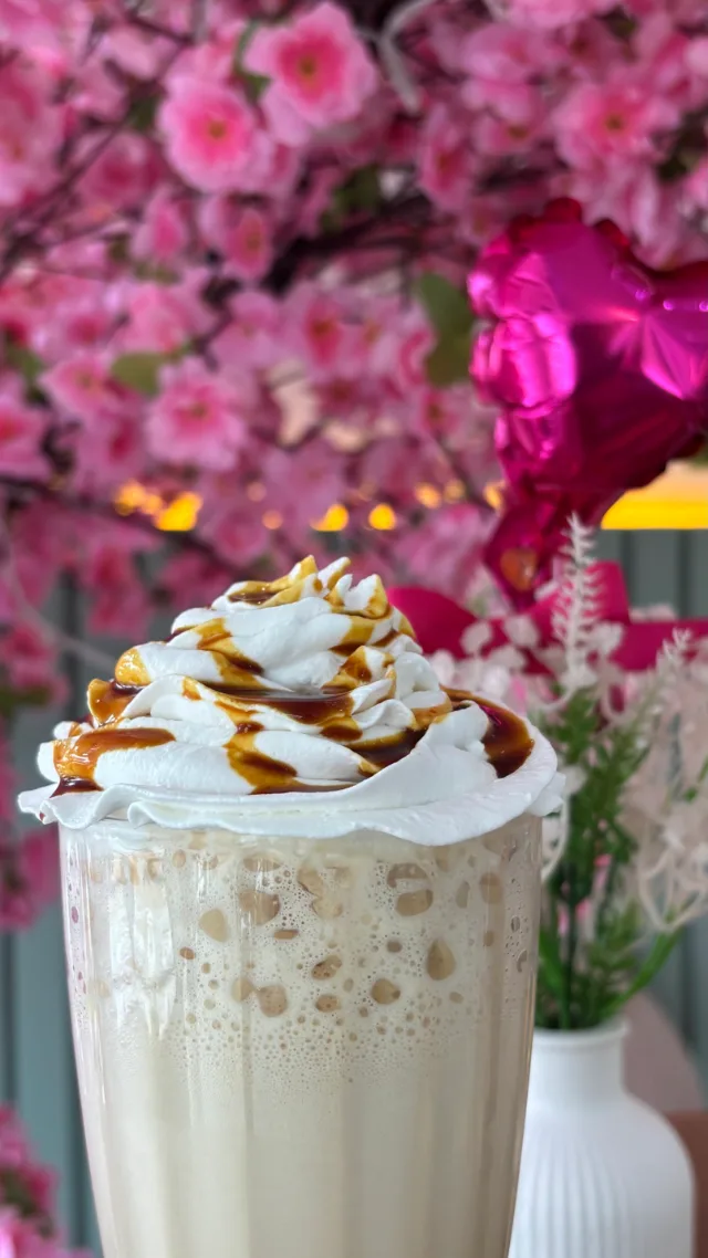 Bebida num copo, coberta de chantilly com fios de calda, com um vaso de flores e a árvore de flores rosa ao fundo.