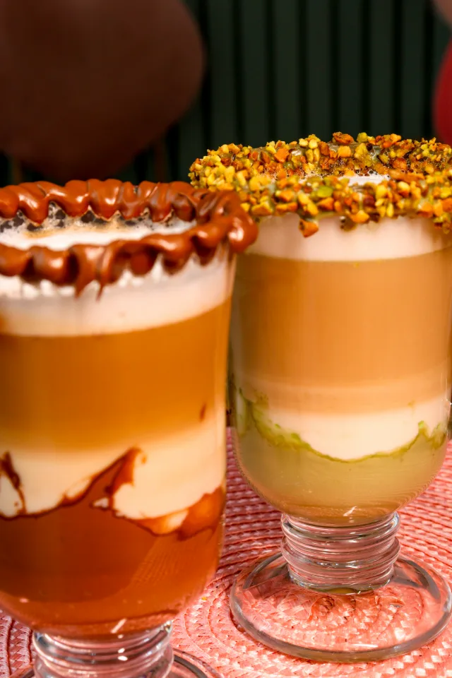 Dois cafés em camadas em canecas de vidro, um com a borda de chocolate e outro com a borda de pistache.