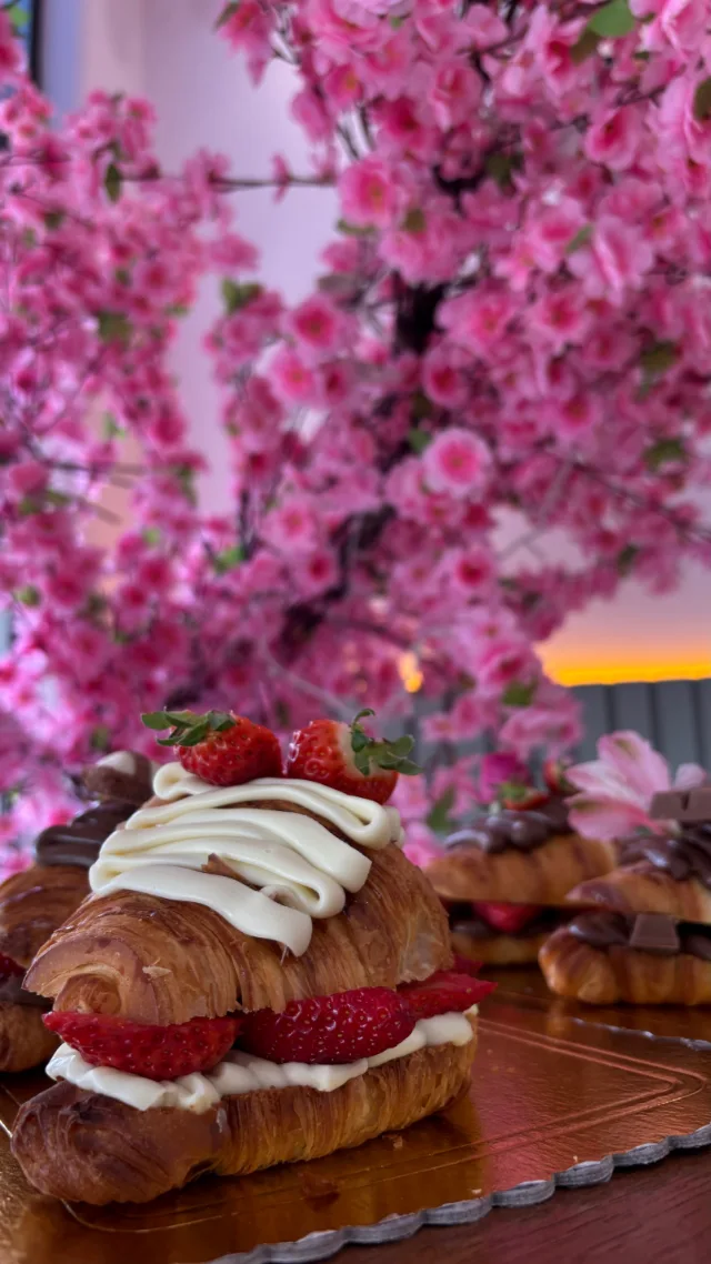 Croissant recheado com creme branco e morangos cortados, com mais creme em ondas e dois morangos inteiros por cima, numa base dourada, diante da árvore de flores rosa.