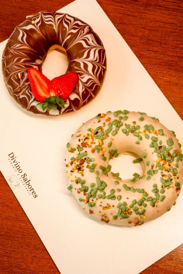 Dois donuts sobre o papel com o nome Divino Sabores: um com cobertura de chocolate marmorizada e morango no meio, outro com cobertura branca e confeitos verdes.