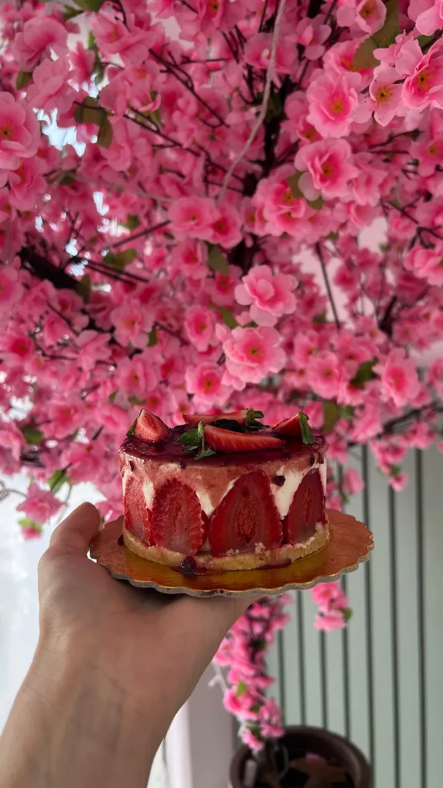 Bolo de mousse cercado de morangos cortados ao meio, com frutas vermelhas por cima, numa base dourada segurada na mão, diante da árvore de flores rosa.