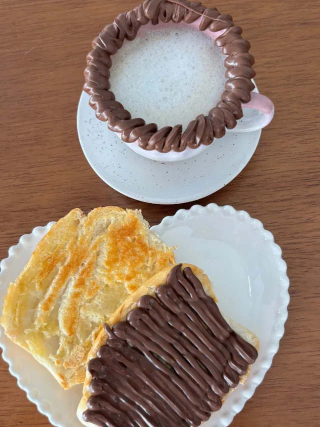 Xícara de café com espuma e a borda de chocolate, e dois crepes dobrados num prato em forma de coração, um com açúcar queimado e outro com fios de chocolate.
