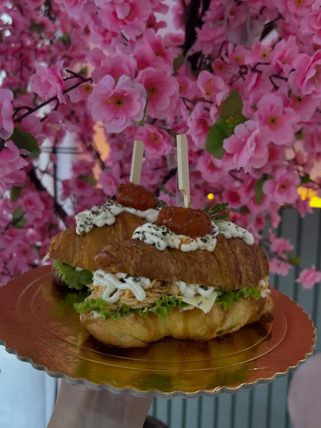 Croissants recheados com frango desfiado, alface e creme branco, com tomatinho no palito por cima, num prato dourado, diante da árvore de flores rosa.