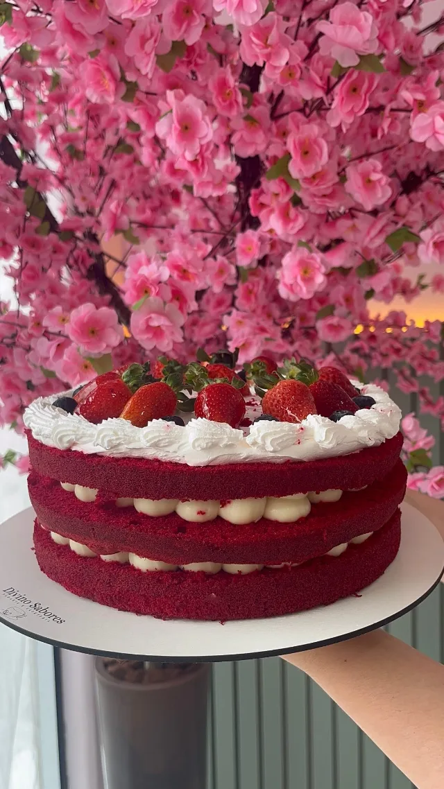 Bolo de massa vermelha em três camadas com recheio branco, coberto de chantilly e morangos, segurado diante da árvore de flores rosa.