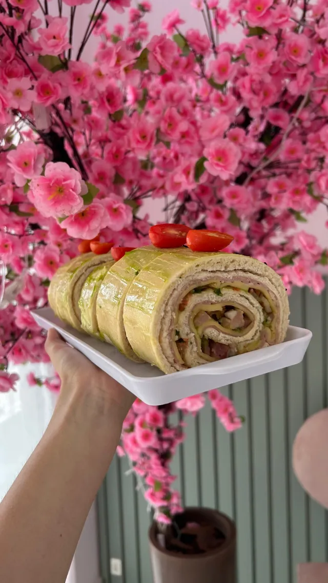 Rocambole recheado, cortado em fatias, com tomatinhos por cima, numa travessa branca segurada diante da árvore de flores rosa.