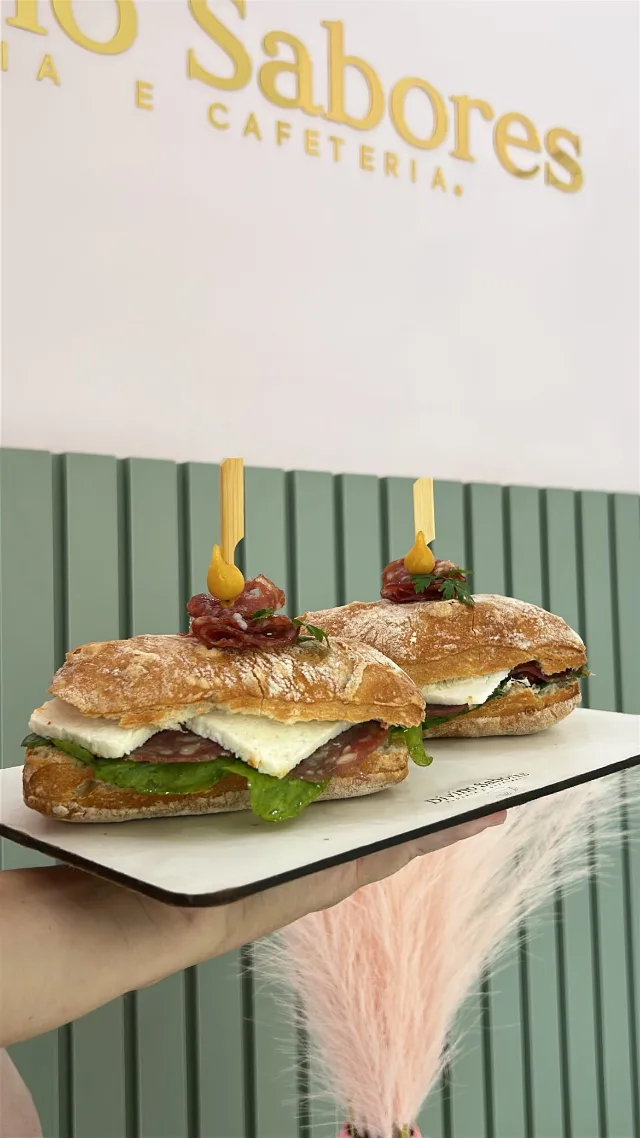 Dois sanduíches no pão rústico com salame, queijo branco e alface, numa tábua branca, diante do painel ripado verde do salão.