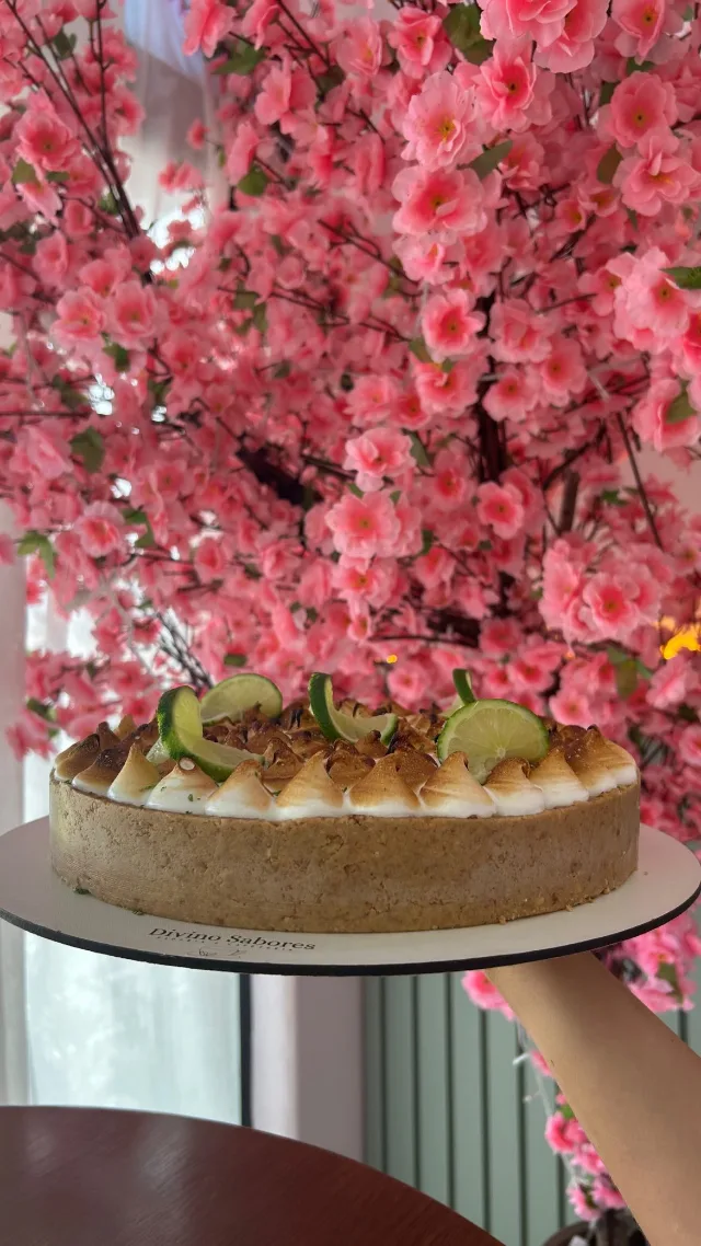 Torta com merengue tostado e rodelas de limão por cima, num prato, segurada diante da árvore de flores rosa.