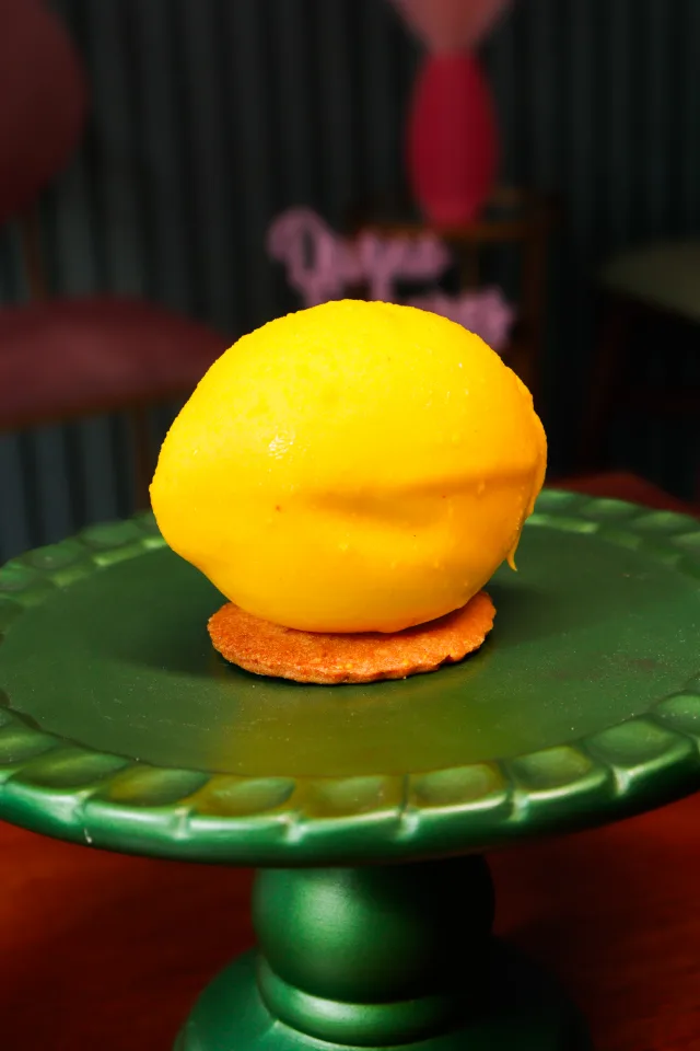 Doce em forma de limão, amarelo, sobre um biscoito, numa boleira verde, com o nome Divino Sabores ao fundo.