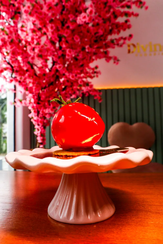 Doce em forma de morango, vermelho e brilhante, numa boleira rosa, com a árvore florida ao fundo.