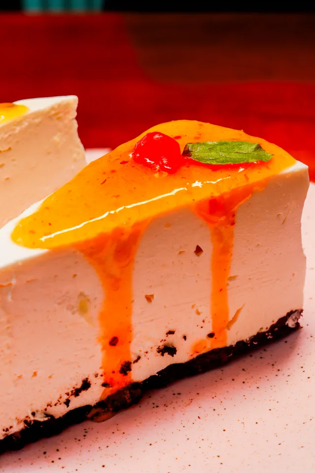 Fatia de torta de creme sobre base escura, com calda de maracujá escorrendo pela lateral, uma cereja e uma folha de hortelã por cima.