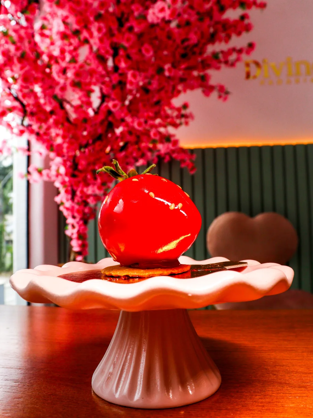 Doce em forma de morango, vermelho e brilhante, numa boleira rosa, com a árvore florida ao fundo.