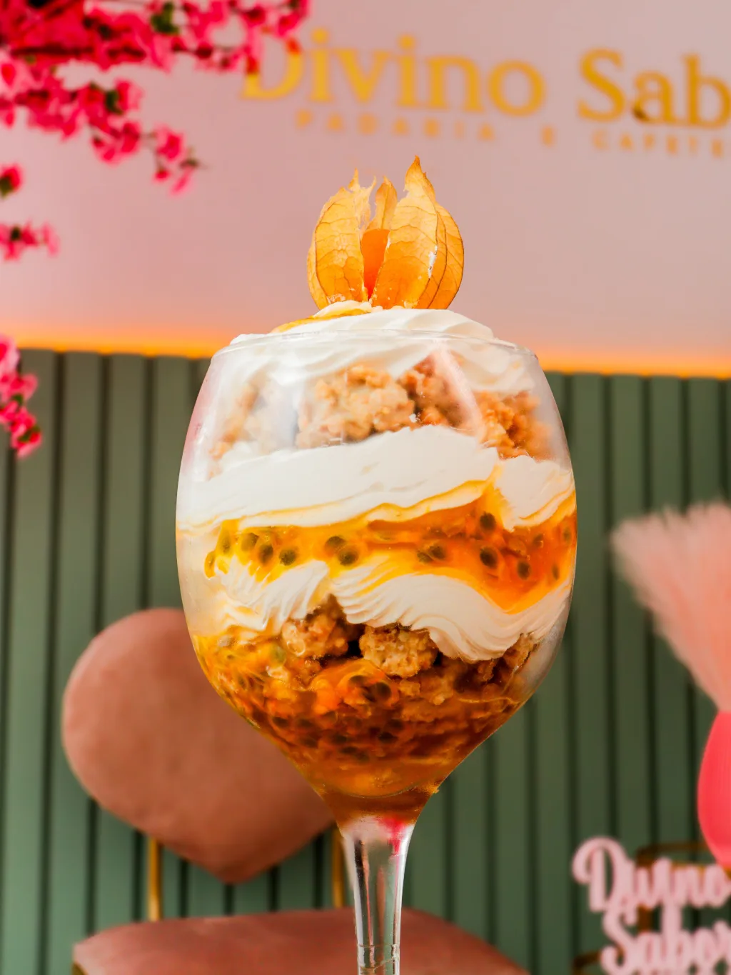 Taça de sobremesa em camadas, com creme e calda de maracujá, decorada com physalis, diante do nome Divino Sabores na parede.